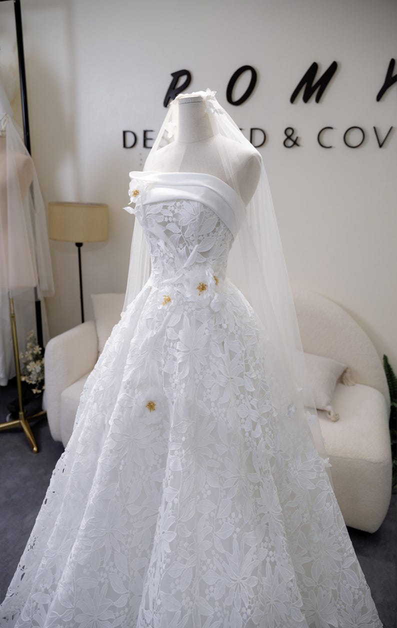 May include: A white bridal gown on a mannequin, with a strapless design and floral lace detailing. The dress has a full skirt and is decorated with small gold accents. A sheer veil is draped from the mannequin's head. The background includes the text "POMH DE D & COV".