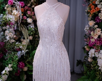 May include: A white sleeveless mini dress featuring a high halter neckline, intricate silver sequin embellishments, and long beaded fringe detailing that hangs from the hem. The garment is displayed on a mannequin with a gold crown topper.