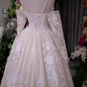 K&ouml;nnte beinhalten: Cremefarbenes Hochzeitskleid auf einer Schaufensterpuppe. Das Kleid hat aufwendige Spitzendetails, lange &Auml;rmel und einen Korsettr&uuml;cken. Das Kleid hat einen weiten Rock und zarte Tr&auml;ger. Der Hintergrund umfasst Blumenarrangements.