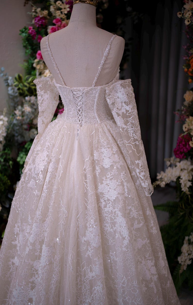 K&ouml;nnte beinhalten: Cremefarbenes Hochzeitskleid auf einer Schaufensterpuppe. Das Kleid hat aufwendige Spitzendetails, lange &Auml;rmel und einen Korsettr&uuml;cken. Das Kleid hat einen weiten Rock und zarte Tr&auml;ger. Der Hintergrund umfasst Blumenarrangements.