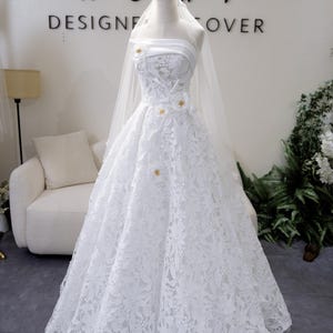 May include: A white bridal gown featuring a floral lace design. The strapless bodice is embellished with small gold details. A sheer veil is draped over the mannequin. The text "ROMY DESIGNER COVER" is visible in the background.