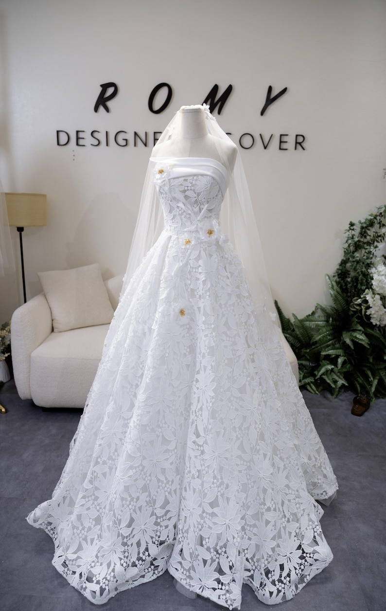 May include: A white bridal gown featuring a floral lace design. The strapless bodice is embellished with small gold details. A sheer veil is draped over the mannequin. The text "ROMY DESIGNER COVER" is visible in the background.