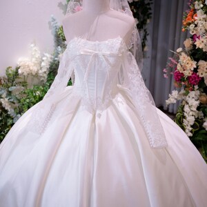 May include: A white ball gown with a corset-style bodice, long lace sleeves, and a full satin skirt. The dress features a sweetheart neckline with a small bow, a sheer veil, and a gold-toned wire crown resting on the mannequin's head.
