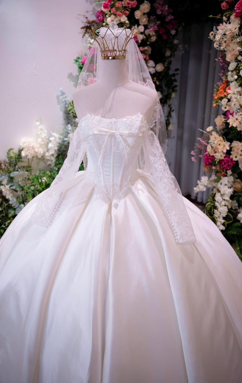 May include: A white ball gown with a corset-style bodice, long lace sleeves, and a full satin skirt. The dress features a sweetheart neckline with a small bow, a sheer veil, and a gold-toned wire crown resting on the mannequin's head.
