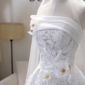 May include: A white wedding dress with a strapless neckline and floral lace detailing. The dress features decorative floral accents with gold embellishments. A sheer veil is draped over the dress form.