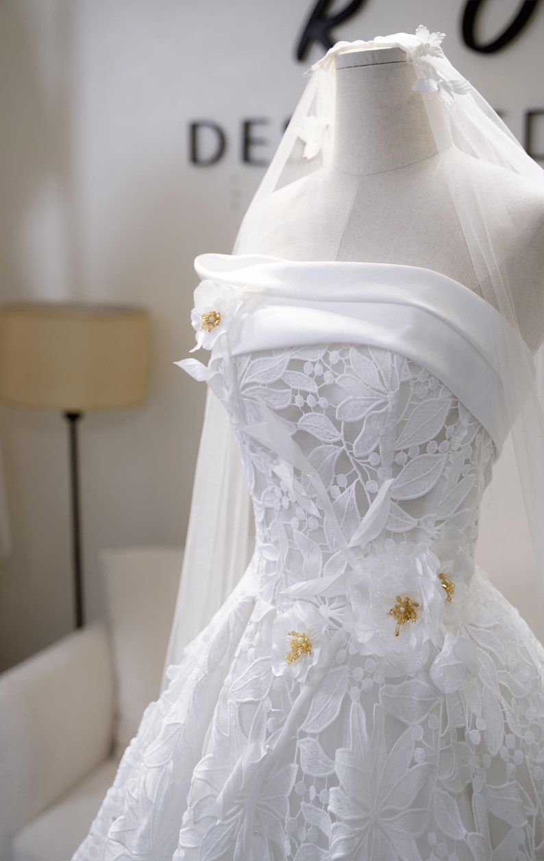 May include: A white wedding dress with a strapless neckline and floral lace detailing. The dress features decorative floral accents with gold embellishments. A sheer veil is draped over the dress form.