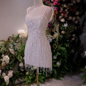May include: A white, beaded mini dress on a mannequin, featuring a fringe hem and spaghetti straps. A gold crown sits atop the mannequin's head. The dress is displayed on a gold stand, with a floral backdrop.