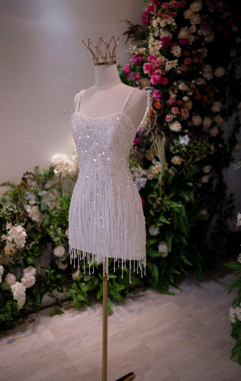 May include: A white, beaded mini dress on a mannequin, featuring a fringe hem and spaghetti straps. A gold crown sits atop the mannequin's head. The dress is displayed on a gold stand, with a floral backdrop.