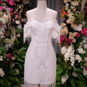 May include: A white off-the-shoulder mini dress featuring intricate silver beadwork, sequin embellishments, and long fringe detailing along the neckline and hem. The garment is displayed on a mannequin topped with a gold wire crown accessory.