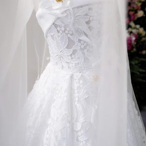 May include: A white wedding dress on a mannequin. The strapless bodice features intricate floral lace, and a delicate veil drapes over the dress. A white flower with gold accents adorns the bodice. The skirt is full and flowing.