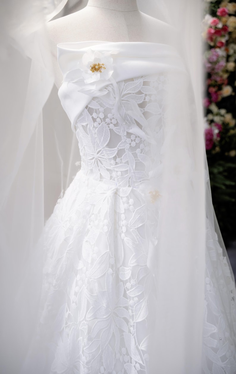 May include: A white wedding dress on a mannequin. The strapless bodice features intricate floral lace, and a delicate veil drapes over the dress. A white flower with gold accents adorns the bodice. The skirt is full and flowing.