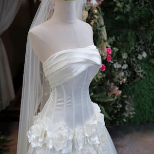 May include: A strapless white wedding dress featuring a structured corset bodice with horizontal pleats, a waistline adorned with large three-dimensional white fabric roses, and a long sheer veil topped with a gold wire crown on a mannequin.