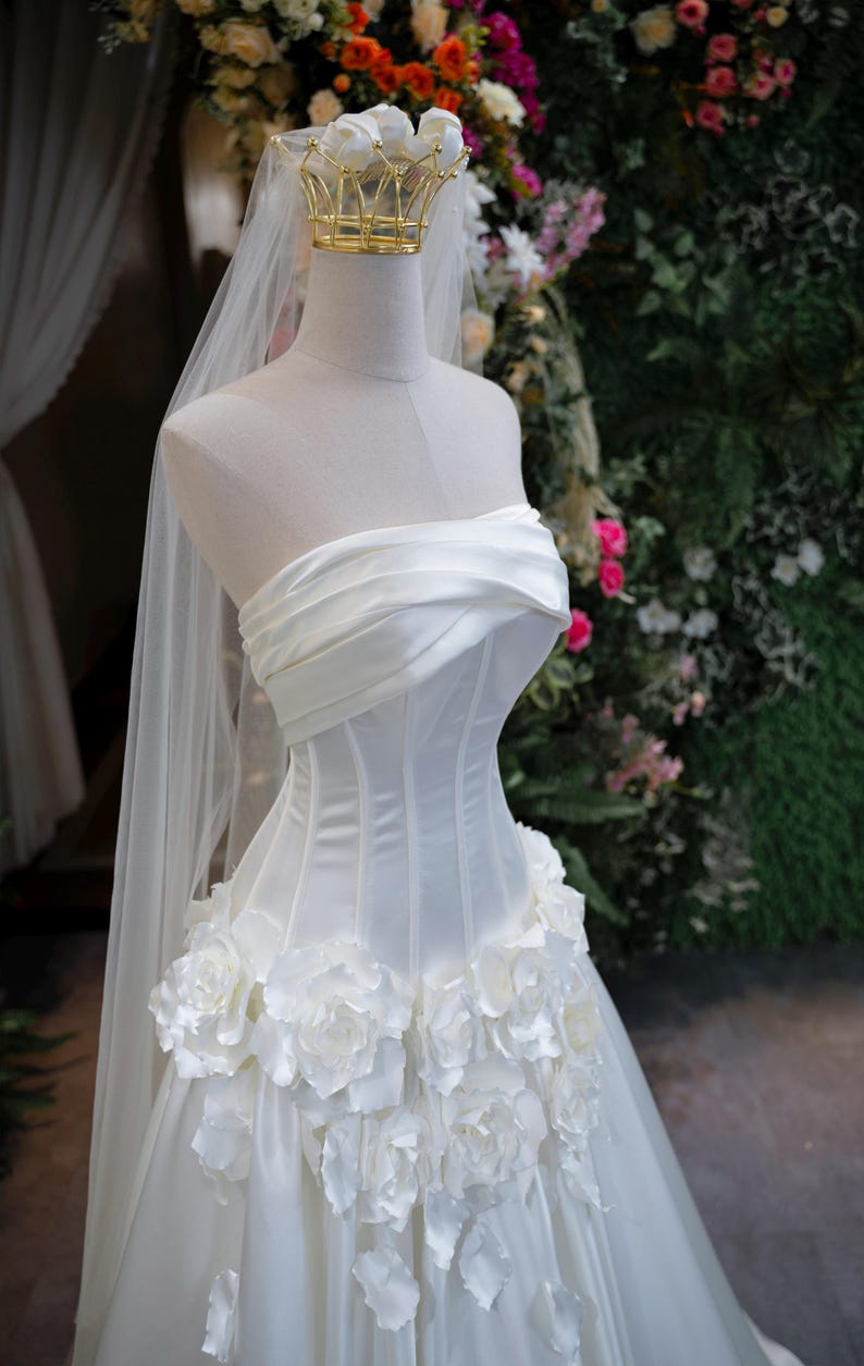 May include: A strapless white wedding dress featuring a structured corset bodice with horizontal pleats, a waistline adorned with large three-dimensional white fabric roses, and a long sheer veil topped with a gold wire crown on a mannequin.