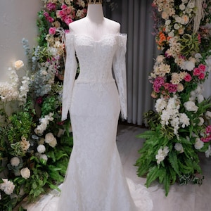 May include: A white, off-the-shoulder wedding dress with long sleeves and a mermaid silhouette. The dress is made of lace and features a sweetheart neckline. A gold crown sits atop the mannequin head. The dress is displayed in front of a floral arch.