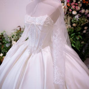 May include: A white ball gown with a corset-style bodice featuring lace overlays and a central ribbon bow. The dress has long, sheer lace sleeves, a voluminous satin skirt, and is paired with a sheer veil and a gold-toned wire crown headpiece.