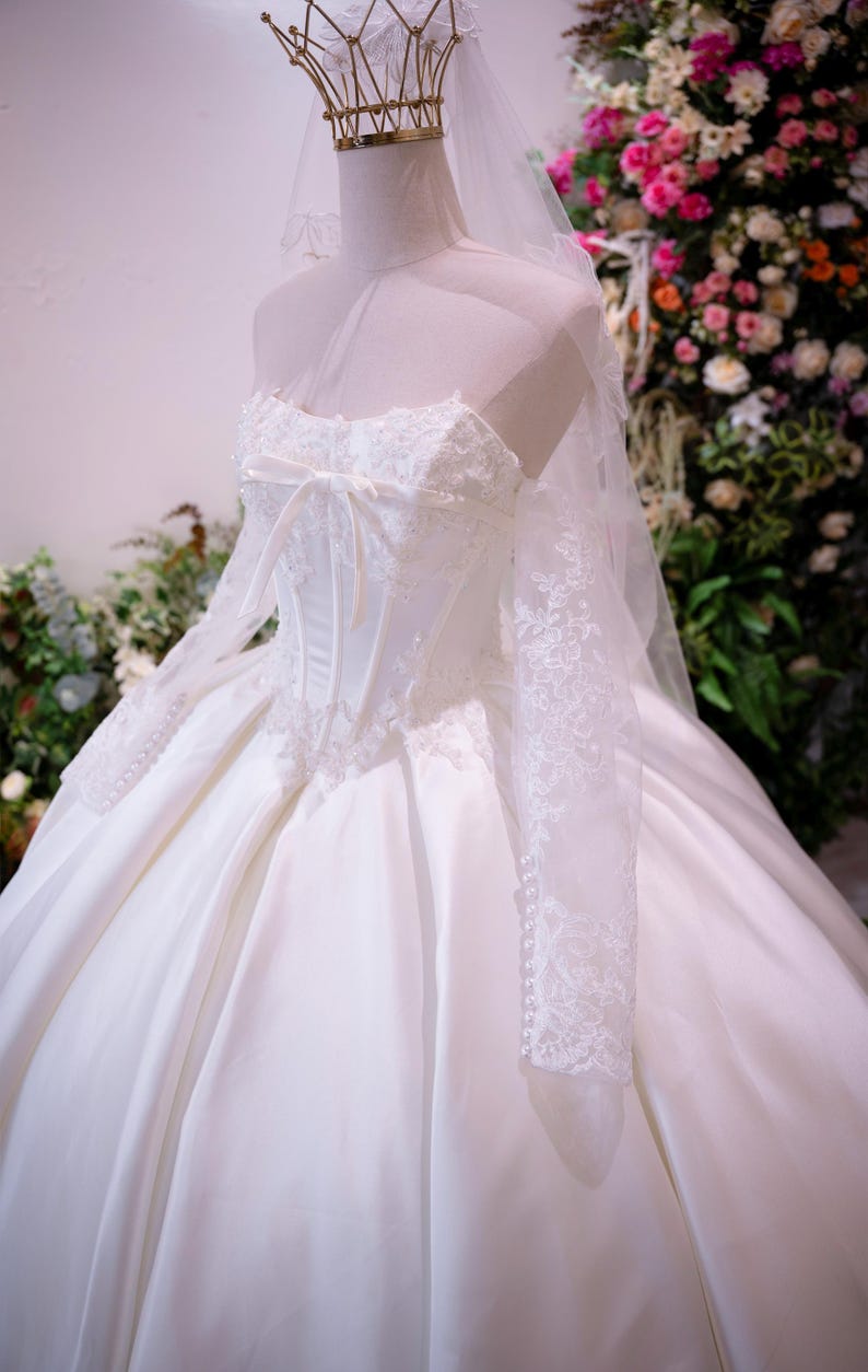May include: A white ball gown with a corset-style bodice featuring lace overlays and a central ribbon bow. The dress has long, sheer lace sleeves, a voluminous satin skirt, and is paired with a sheer veil and a gold-toned wire crown headpiece.