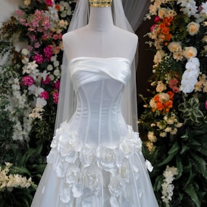 May include: A white strapless corset-style gown featuring a draped bodice and a skirt adorned with three-dimensional white fabric roses. A sheer white veil is attached to a gold-toned crown headpiece resting atop the mannequin's neck.