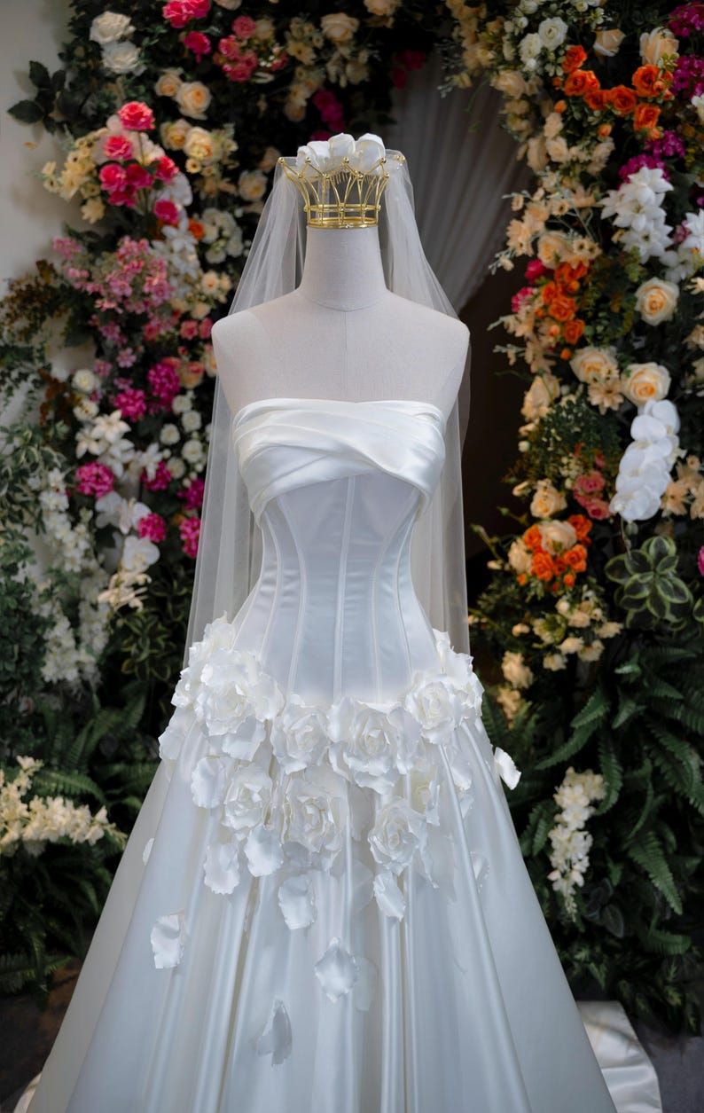 May include: A white strapless corset-style gown featuring a draped bodice and a skirt adorned with three-dimensional white fabric roses. A sheer white veil is attached to a gold-toned crown headpiece resting atop the mannequin's neck.