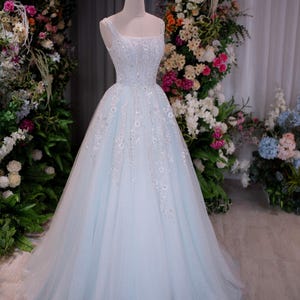 May include: A light blue, one-shoulder formal gown with a full skirt. The bodice is embellished with sequins and floral appliques. A gold crown sits atop the mannequin head. The dress is displayed in front of a floral arch.