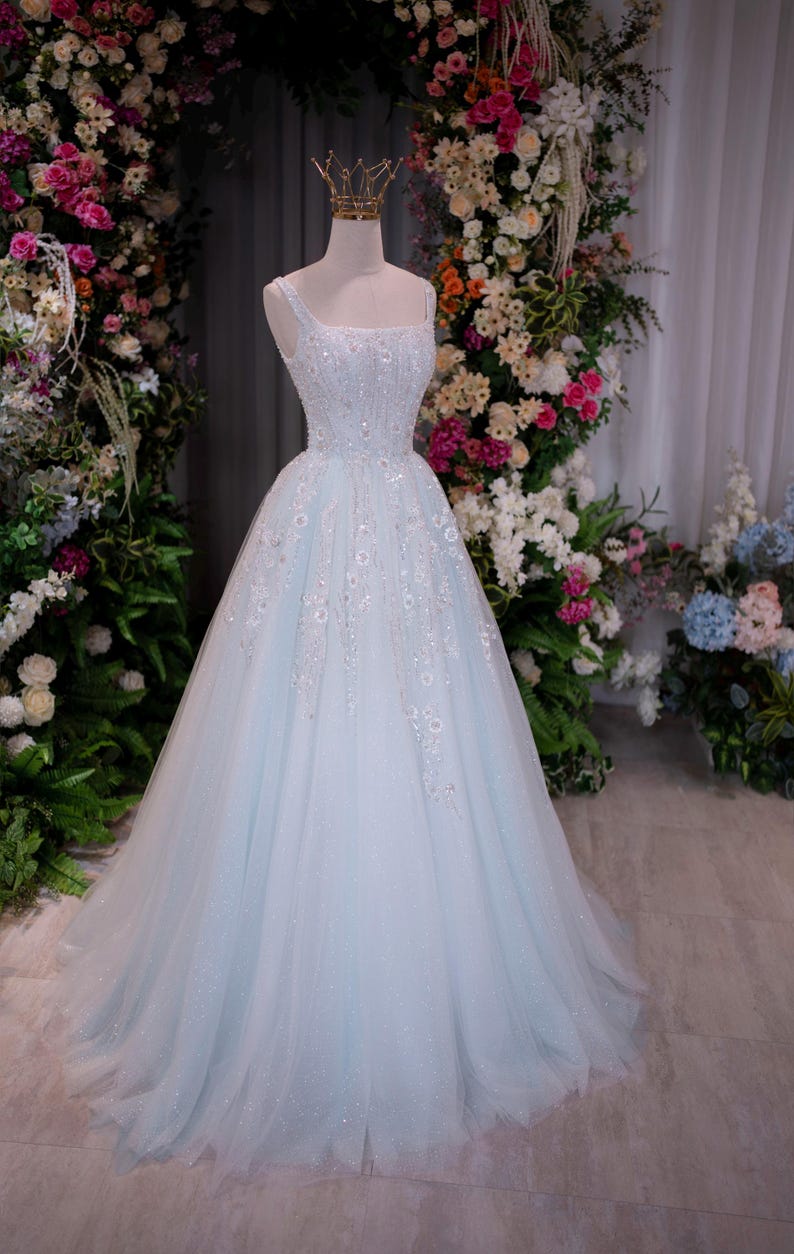 May include: A light blue, one-shoulder formal gown with a full skirt. The bodice is embellished with sequins and floral appliques. A gold crown sits atop the mannequin head. The dress is displayed in front of a floral arch.