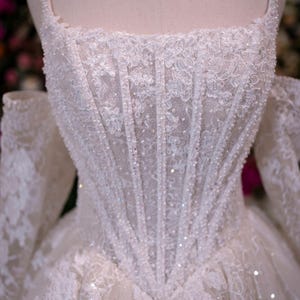 K&ouml;nnte beinhalten: Wei&szlig;es Hochzeitskleid mit quadratischem Ausschnitt und schulterfreien &Auml;rmeln. Das Oberteil ist aufwendig mit Spitze verziert und mit glitzernden Perlen besetzt. Der Rock verl&auml;uft von der Taille aus und bildet eine klassische Silhouette.