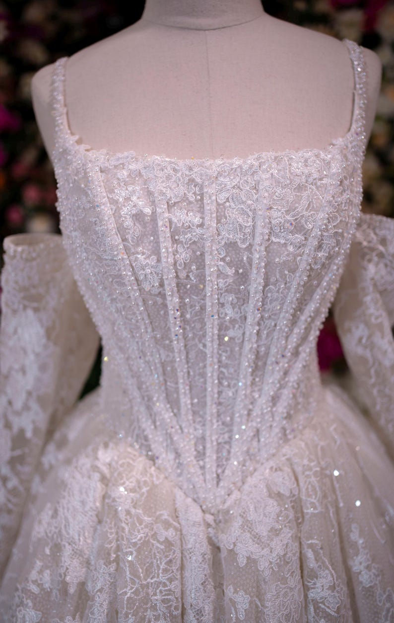 K&ouml;nnte beinhalten: Wei&szlig;es Hochzeitskleid mit quadratischem Ausschnitt und schulterfreien &Auml;rmeln. Das Oberteil ist aufwendig mit Spitze verziert und mit glitzernden Perlen besetzt. Der Rock verl&auml;uft von der Taille aus und bildet eine klassische Silhouette.