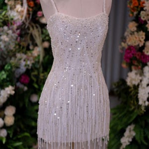 May include: A white, beaded mini dress on a mannequin. The dress features a square neckline, spaghetti straps, and a fitted bodice. The skirt is adorned with vertical beaded fringe. The dress is embellished with sequins and small beads.