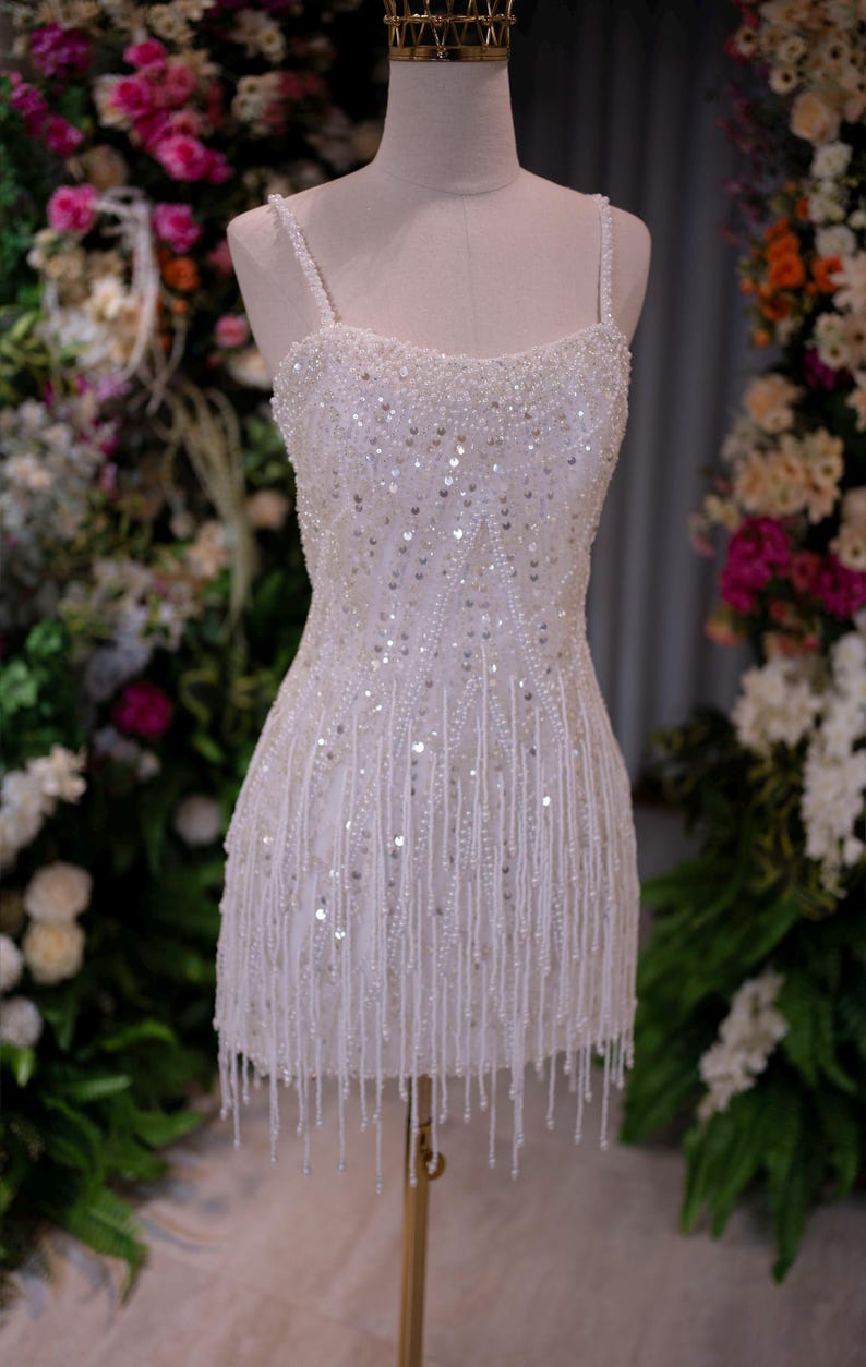 May include: A white, beaded mini dress on a mannequin. The dress features a square neckline, spaghetti straps, and a fitted bodice. The skirt is adorned with vertical beaded fringe. The dress is embellished with sequins and small beads.