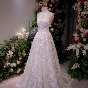 May include: A floor-length white lace gown featuring a high halter neckline with sheer illusion detailing, a fitted bodice adorned with intricate floral embroidery, and a full A-line skirt that extends into a train, displayed on a mannequin.