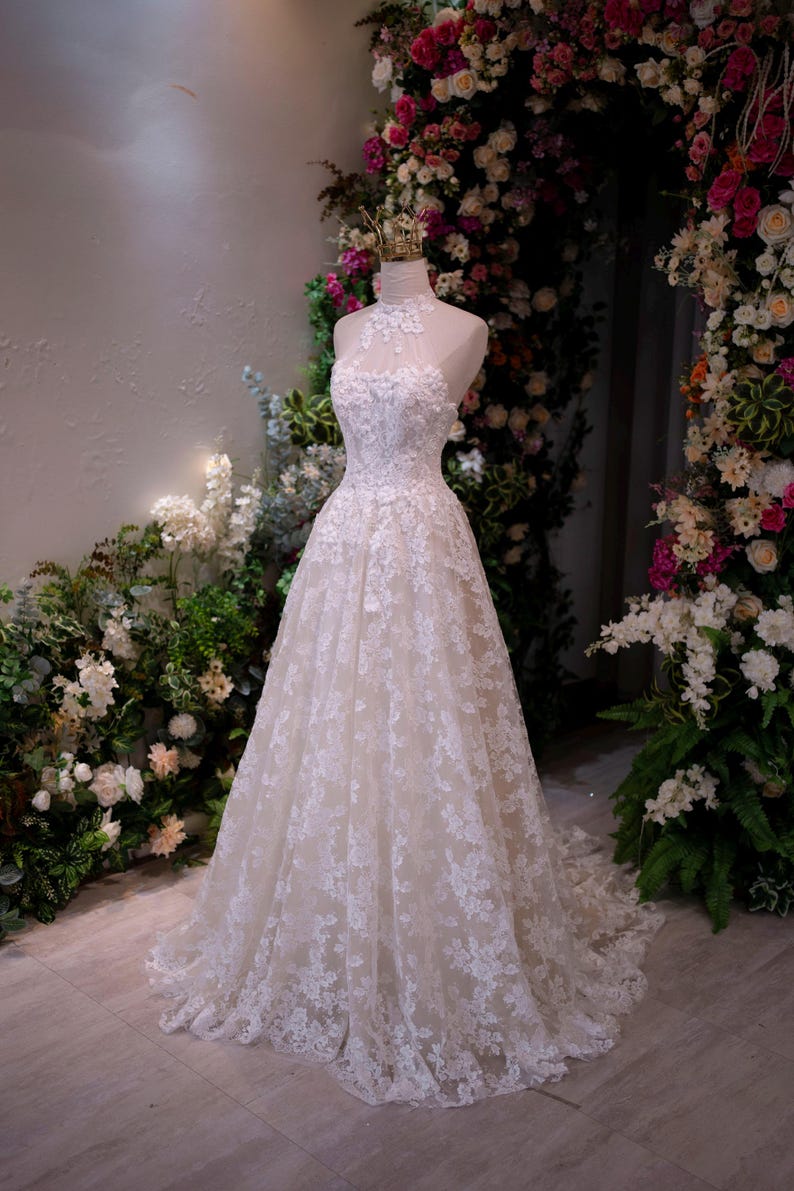 May include: A floor-length white lace gown featuring a high halter neckline with sheer illusion detailing, a fitted bodice adorned with intricate floral embroidery, and a full A-line skirt that extends into a train, displayed on a mannequin.