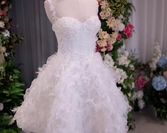 Puede incluir: Un vestido corto blanco sobre un maniquí con escote corazón, cuerpo con cuentas y tirantes finos. La falda está confeccionada con capas de pétalos de tela transparentes y volantes, junto con plumas, creando un aspecto voluminoso y texturizado.