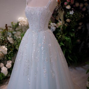May include: A light blue wedding dress with a square neckline and spaghetti straps. The bodice and skirt are embellished with sequins and floral accents. A gold crown sits atop the mannequin's head. The dress is displayed in front of a floral backdrop.