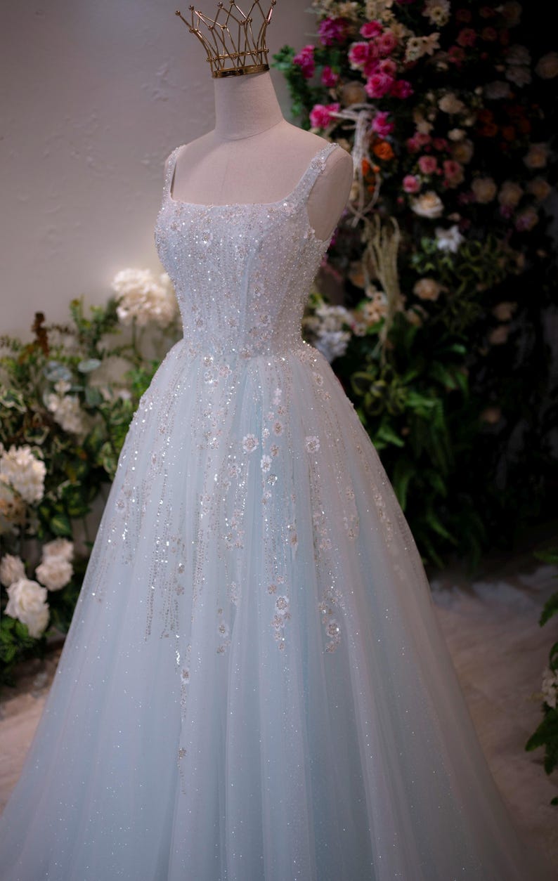 May include: A light blue wedding dress with a square neckline and spaghetti straps. The bodice and skirt are embellished with sequins and floral accents. A gold crown sits atop the mannequin's head. The dress is displayed in front of a floral backdrop.