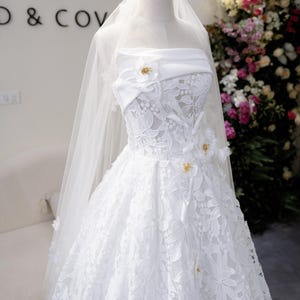 May include: A white strapless wedding dress with floral lace detailing and a flowing skirt. The dress features a sweetheart neckline and is adorned with small, gold-coloured embellishments. A sheer veil drapes over the dress, adding to its elegance.