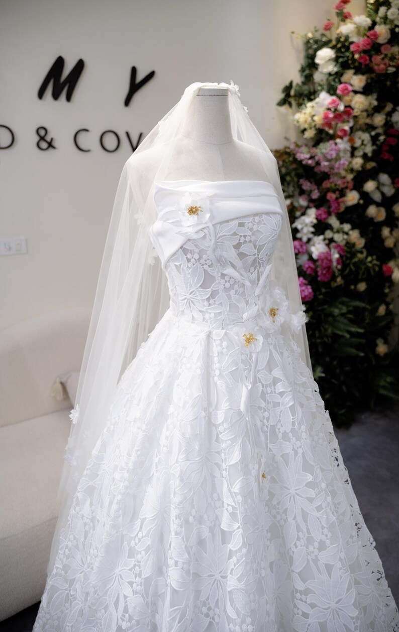 May include: A white strapless wedding dress with floral lace detailing and a flowing skirt. The dress features a sweetheart neckline and is adorned with small, gold-coloured embellishments. A sheer veil drapes over the dress, adding to its elegance.