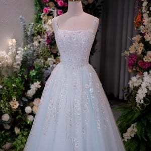 May include: A light blue, floor-length formal gown with a square neckline and embellished bodice. The dress is adorned with sparkling sequins and floral accents. A gold crown sits atop the mannequin head. The background features floral arrangements.