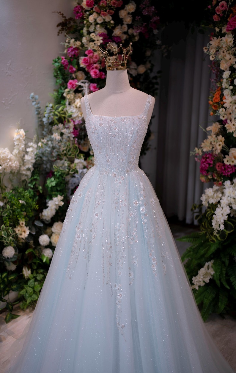 May include: A light blue, floor-length formal gown with a square neckline and embellished bodice. The dress is adorned with sparkling sequins and floral accents. A gold crown sits atop the mannequin head. The background features floral arrangements.