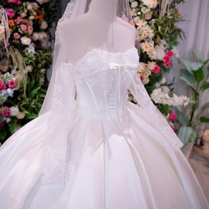 May include: A white strapless ball gown with a corset-style bodice, lace long sleeves, and a full satin skirt. The dress features a small bow at the center neckline, delicate lace appliques, and a sheer veil topped with a gold metal crown.