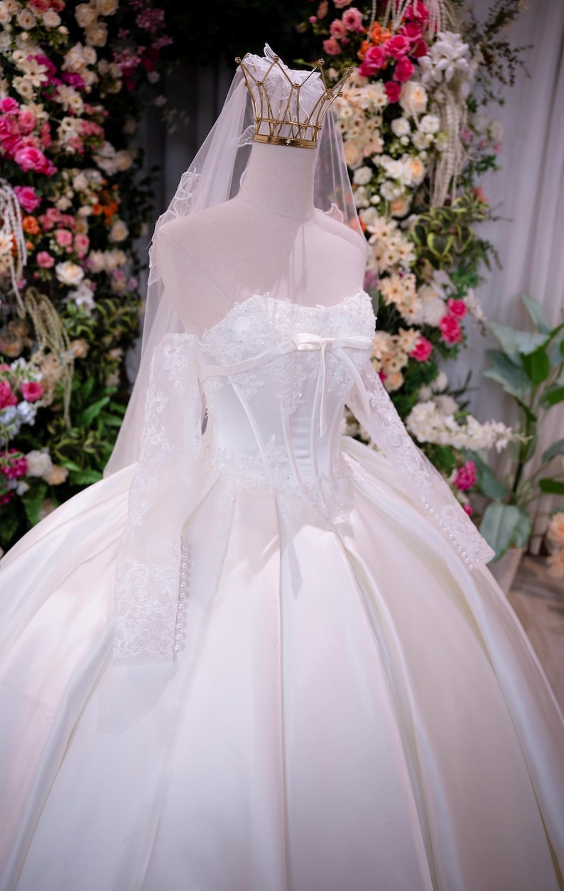 May include: A white strapless ball gown with a corset-style bodice, lace long sleeves, and a full satin skirt. The dress features a small bow at the center neckline, delicate lace appliques, and a sheer veil topped with a gold metal crown.