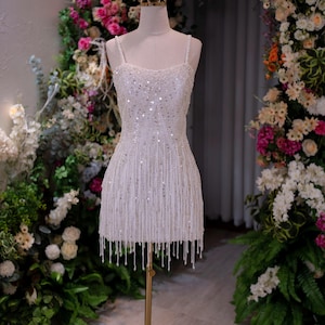 May include: A white, beaded mini dress with spaghetti straps and a fringe hemline. The dress is displayed on a mannequin with a gold crown, set against a backdrop of flowers and greenery. The dress is approximately 30 inches in length.