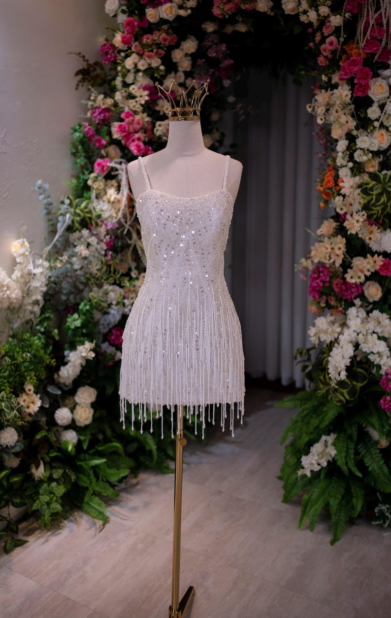 May include: A white, beaded mini dress with spaghetti straps and a fringe hemline. The dress is displayed on a mannequin with a gold crown, set against a backdrop of flowers and greenery. The dress is approximately 30 inches in length.