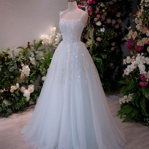 May include: A light blue wedding gown on a mannequin. The dress features a square neckline, embellished bodice, and a flowing tulle skirt. A gold crown sits atop the mannequin's head. The background includes floral arrangements.