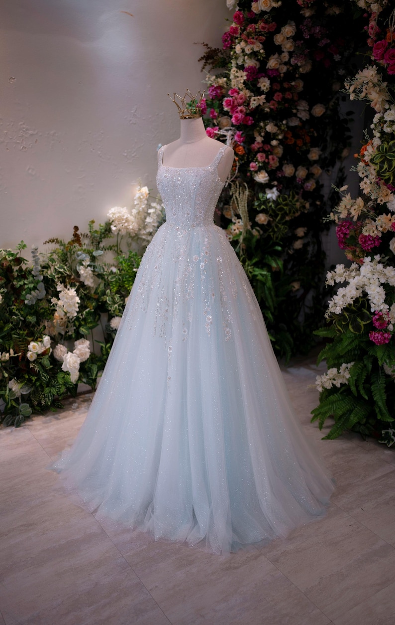 May include: A light blue wedding gown on a mannequin. The dress features a square neckline, embellished bodice, and a flowing tulle skirt. A gold crown sits atop the mannequin's head. The background includes floral arrangements.