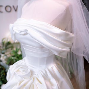 May include: A white satin wedding dress on a mannequin. The strapless bodice features draped fabric, and the full skirt has a ruffled design. A sheer veil and a gold crown are also visible.