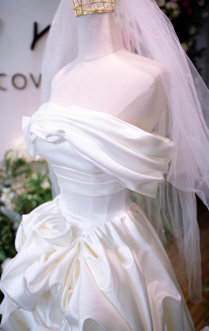 May include: A white satin wedding dress on a mannequin. The strapless bodice features draped fabric, and the full skirt has a ruffled design. A sheer veil and a gold crown are also visible.