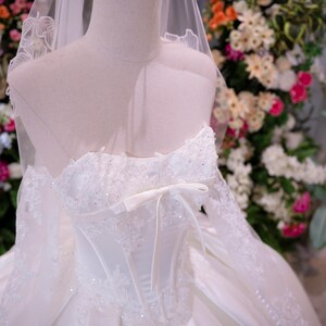May include: A white strapless ball gown featuring a corset-style bodice with intricate lace appliques, sparkling beadwork, and a central ribbon bow. The dress includes long, sheer lace sleeves, a full skirt, and a matching veil with a gold crown.