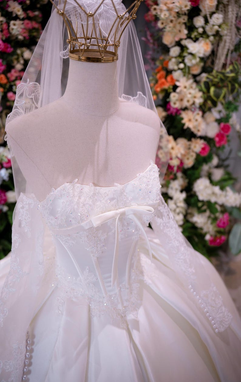 May include: A white strapless ball gown featuring a corset-style bodice with intricate lace appliques, sparkling beadwork, and a central ribbon bow. The dress includes long, sheer lace sleeves, a full skirt, and a matching veil with a gold crown.