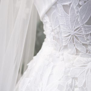 May include: Close-up of a white wedding dress with detailed floral lace. The dress has a structured bodice and a flowing skirt, with a sheer veil on the left. The overall aesthetic is elegant and romantic, perfect for a special occasion.