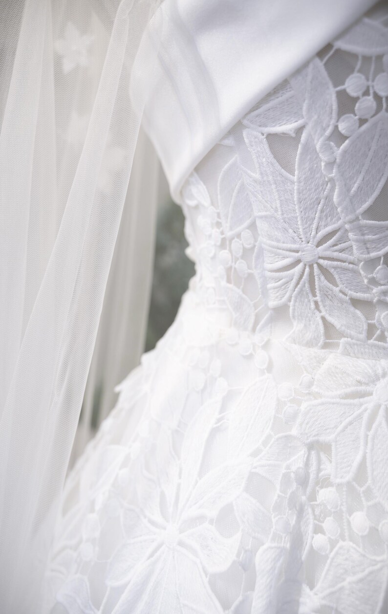 May include: Close-up of a white wedding dress with detailed floral lace. The dress has a structured bodice and a flowing skirt, with a sheer veil on the left. The overall aesthetic is elegant and romantic, perfect for a special occasion.
