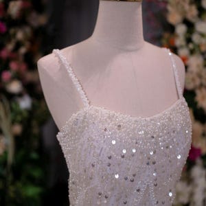 May include: A white, beaded wedding dress with a sweetheart neckline and spaghetti straps. The dress is adorned with sequins and small beads. A gold crown sits atop a mannequin head, suggesting a bridal theme.
